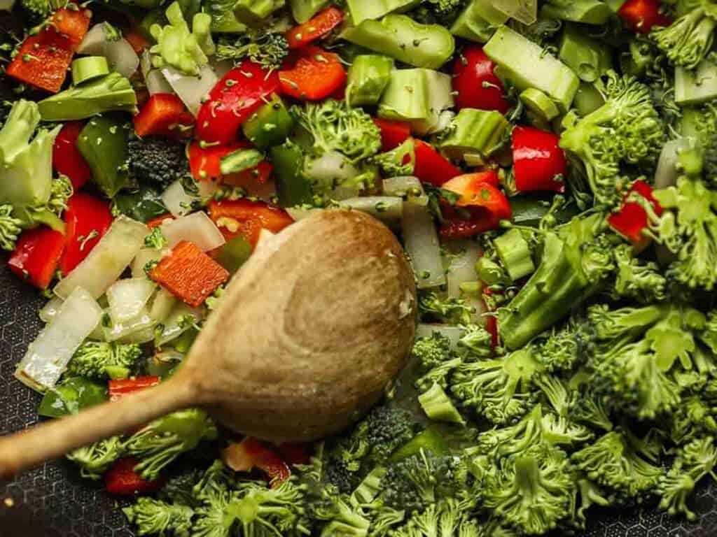 Close-up of chopped broccoli, red bell peppers, onions, and small green vegetables in a pan. A wooden spoon is stirring the colorful mix, suggesting a healthy vegetable stir-fry being prepared.